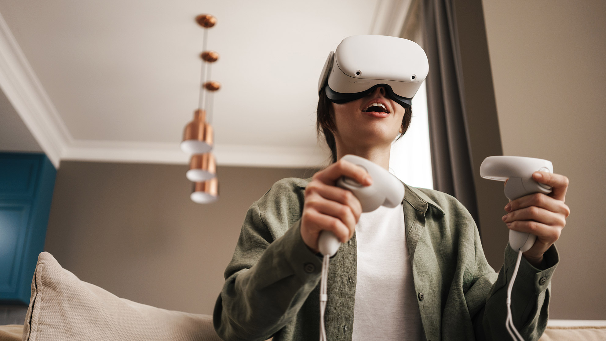 Woman sitting on a beige sofa in a living room, wearing VR glasses and holding game controllers in both hands