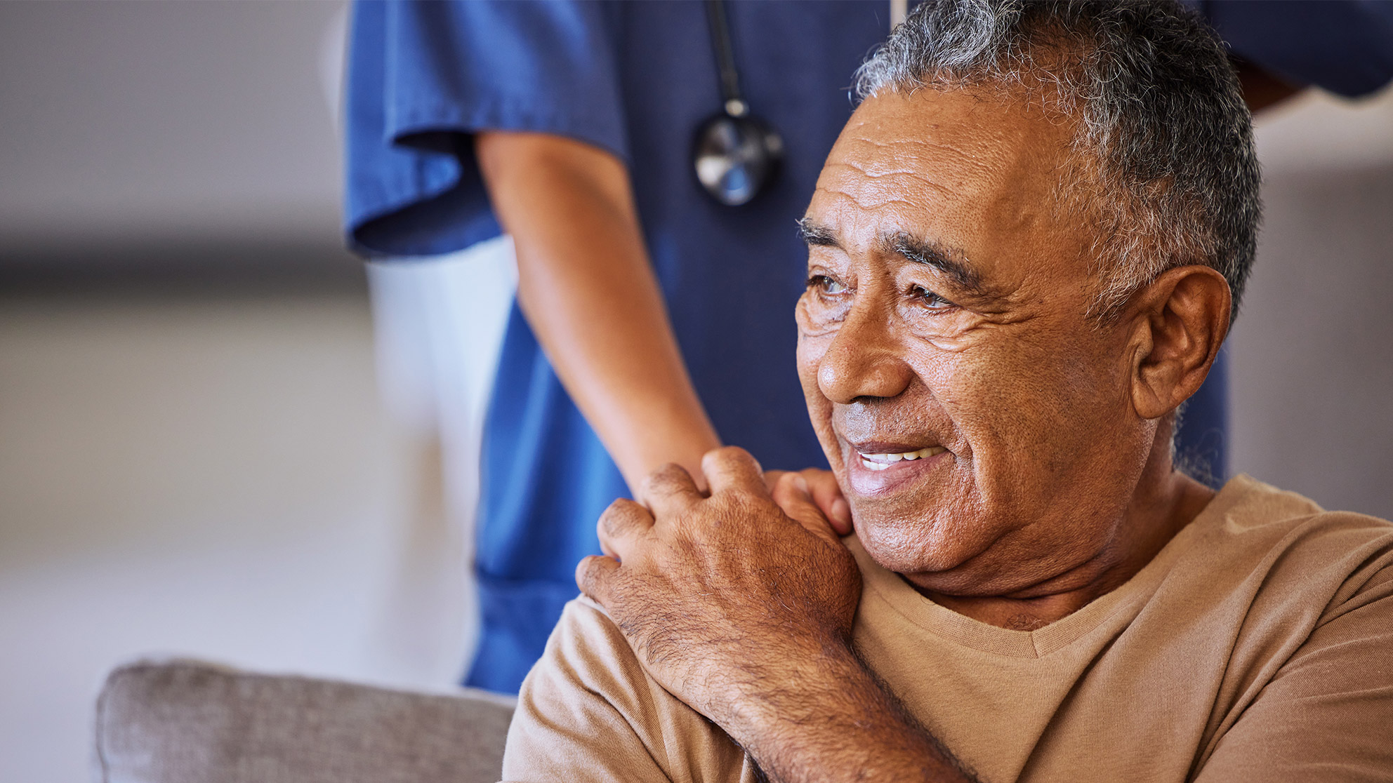 older man sits smiling on a gray sofa and holds the hand of a nurse standing behind him