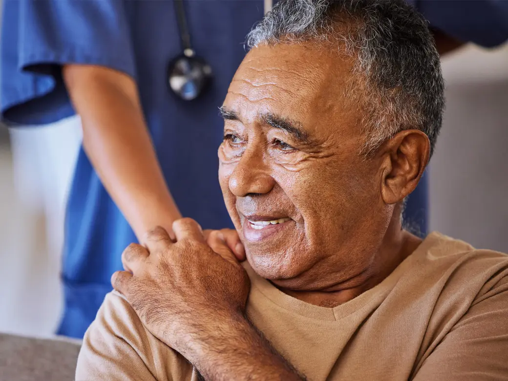 older man sits smiling on a gray sofa and holds the hand of a nurse standing behind him