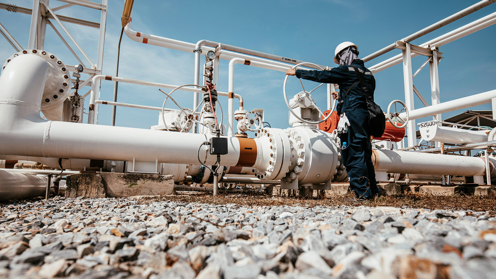 Mechanic turns a large wheel on a pipeline on a platform full of pipes surrounded by stony ground and blue sky