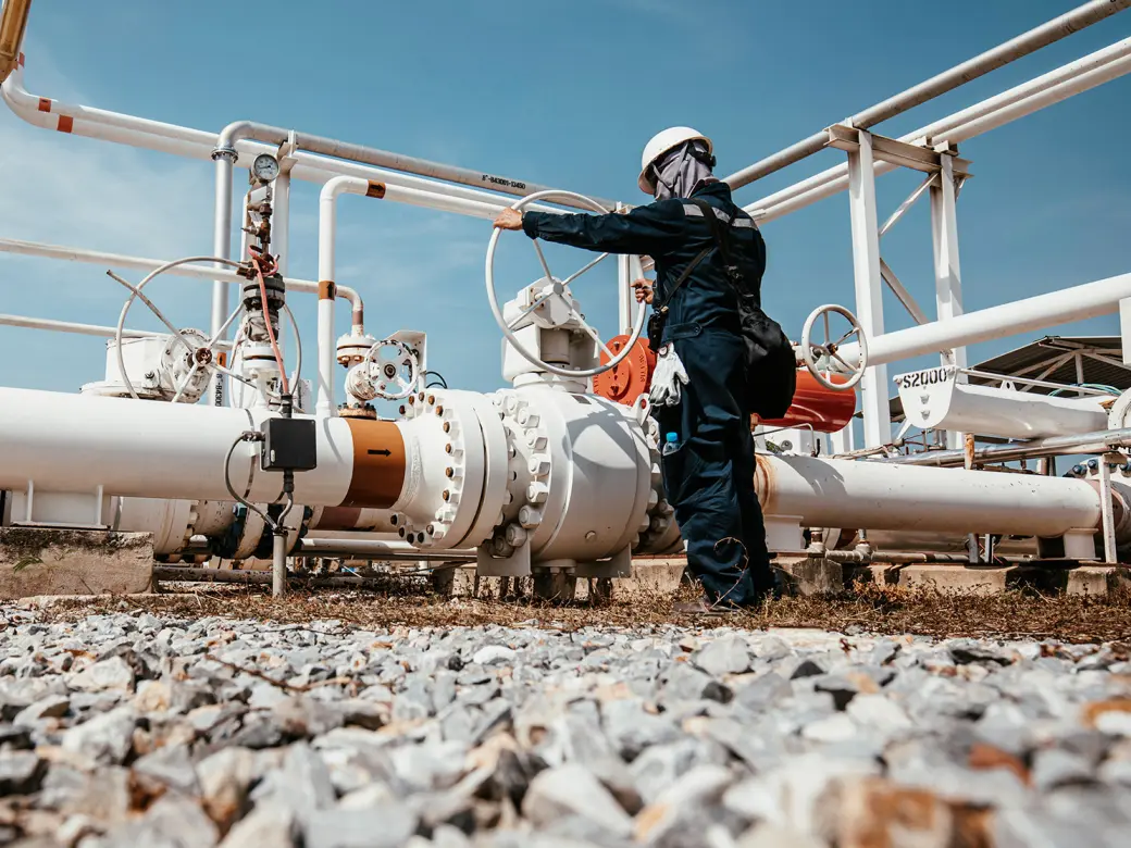 Mechanic turns a large wheel on a pipeline on a platform full of pipes surrounded by stony ground and blue sky