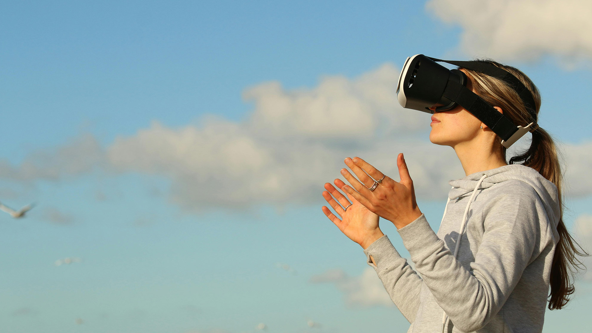 Woman in front of sky with seagull in the background, wearing VR glasses and spreading out her hands